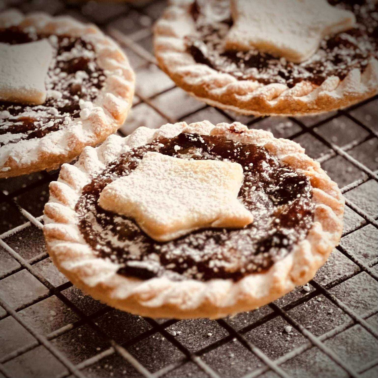 Three Brandy Laced Luxury Mince Pies by Mud Pies, with crimped edges and rich filling, topped with sugared star pastry on a rack.