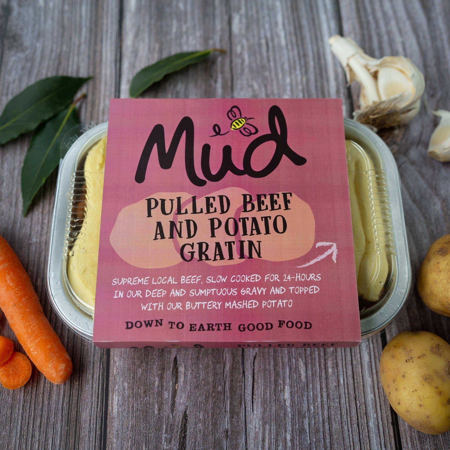 Mud Pies Pulled Beef and Potato Gratin frozen meal on a wooden table, with carrots, garlic and potatoes; beef in rich gravy, buttery mash.