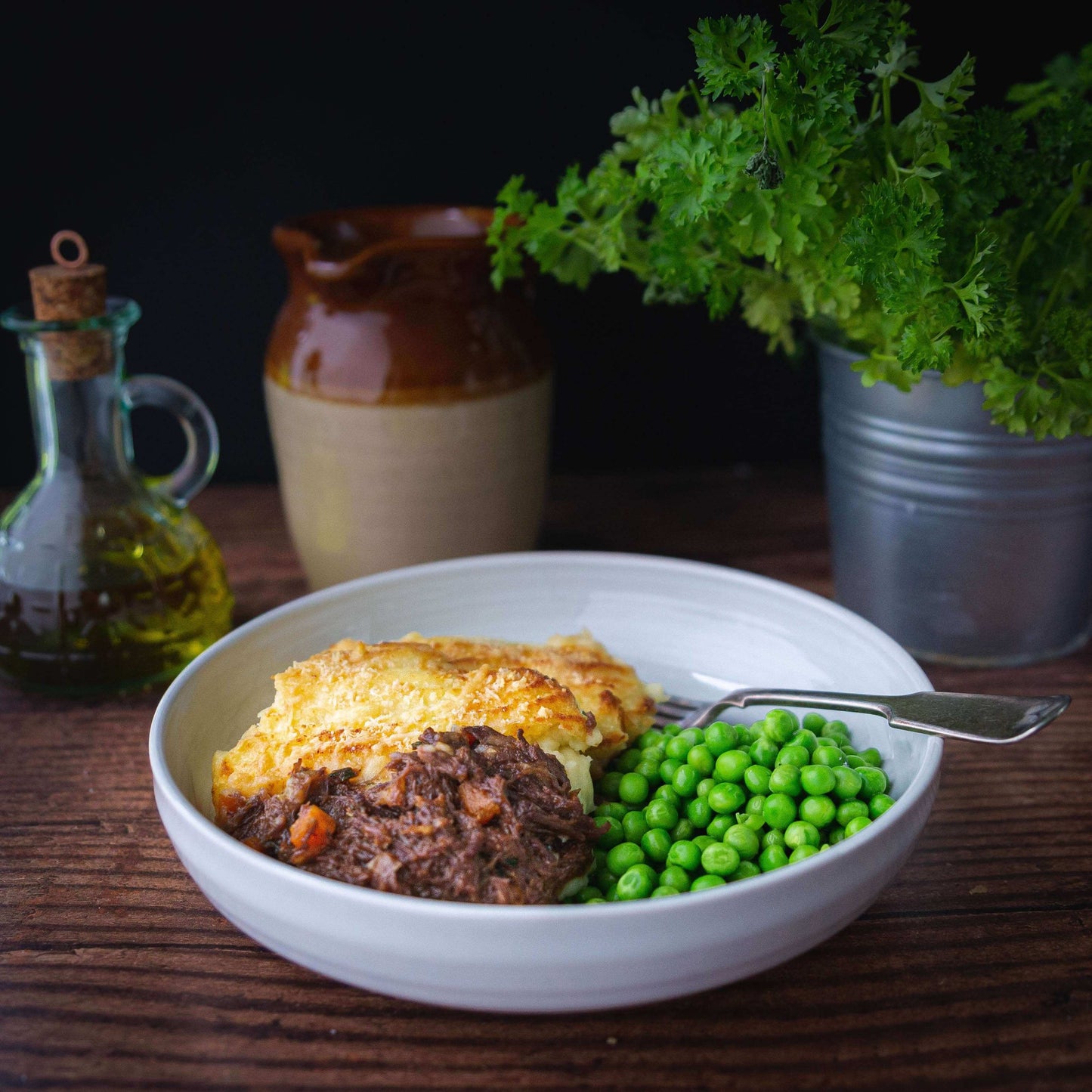 Mud Pies Pulled Beef and Potato Gratin served with green peas in a white bowl, fork beside it on a wooden table; olive oil, jug and parsley behind.