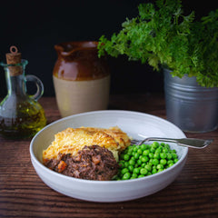 Mud Pies Pulled Beef and Potato Gratin served with green peas in a white bowl, fork beside it on a wooden table; olive oil, jug and parsley behind.