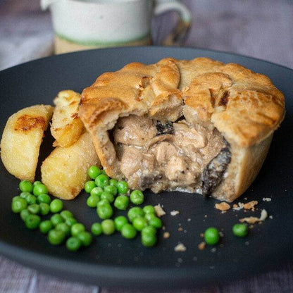 Mud Pies’ Chicken and Mushroom Pie on a black plate, golden pastry sliced to reveal creamy filling, with roast potatoes and peas.