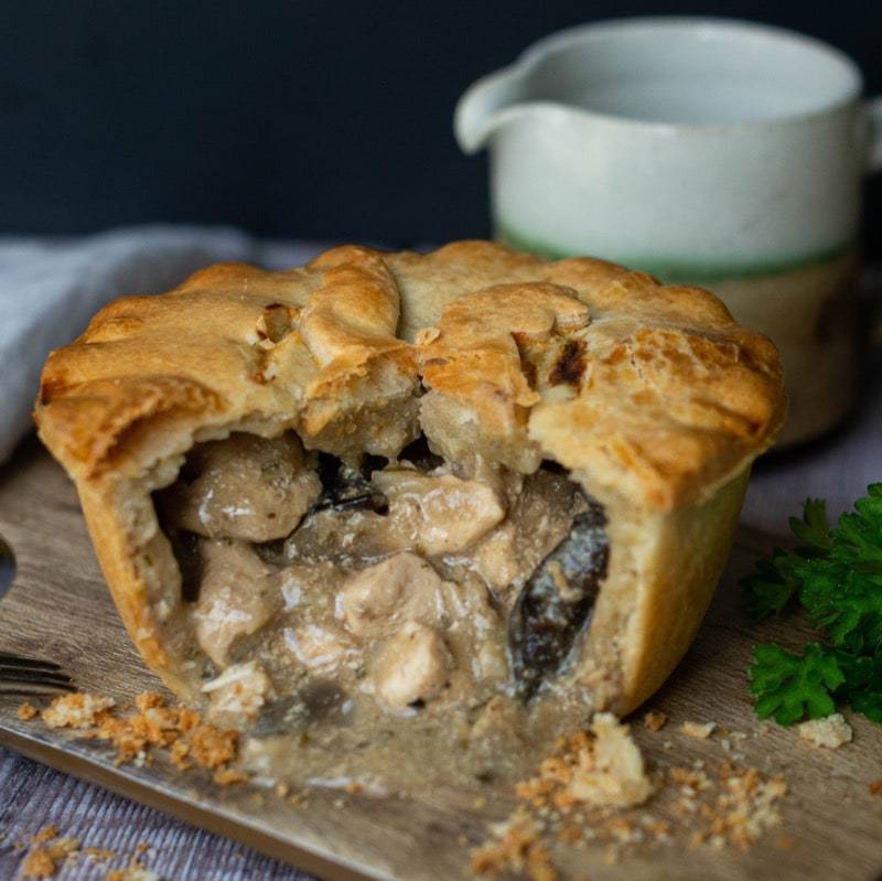 A golden-brown chicken and mushroom pie from Mud Pies is cut open, revealing creamy filling; parsley and crumbs on wooden board.