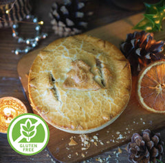 Gluten-Free Christmas In A Pie by Mud Pies, golden-brown with leaf design, sits on wood with pinecones, orange, greenery, candle and gluten-free badge.