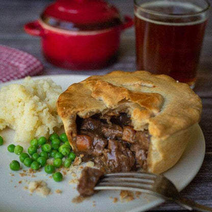 Mud Pies’ Gluten-Free Steak and Ale Pie, sliced to reveal hearty beef and veg, with mash, peas, a fork, dark beer and red pot nearby.