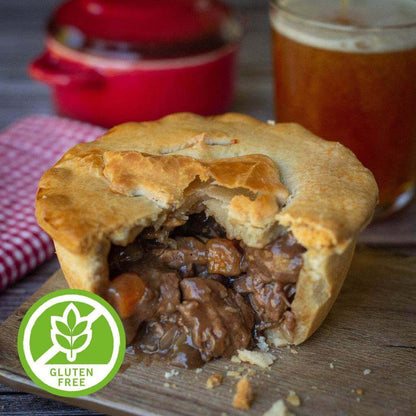 The Mud Pies Gluten-Free Steak and Ale Pie, golden and flaky, sits half-eaten on a wooden board with rich beef filling exposed; an amber beer and red pot rest behind on a checked cloth.