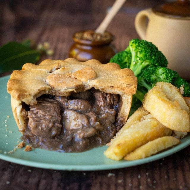 Mud Pies Gluten-Free Steak and Kidney Pie, opened to reveal its rich filling, with golden chips and broccoli; mustard jar and teapot behind.