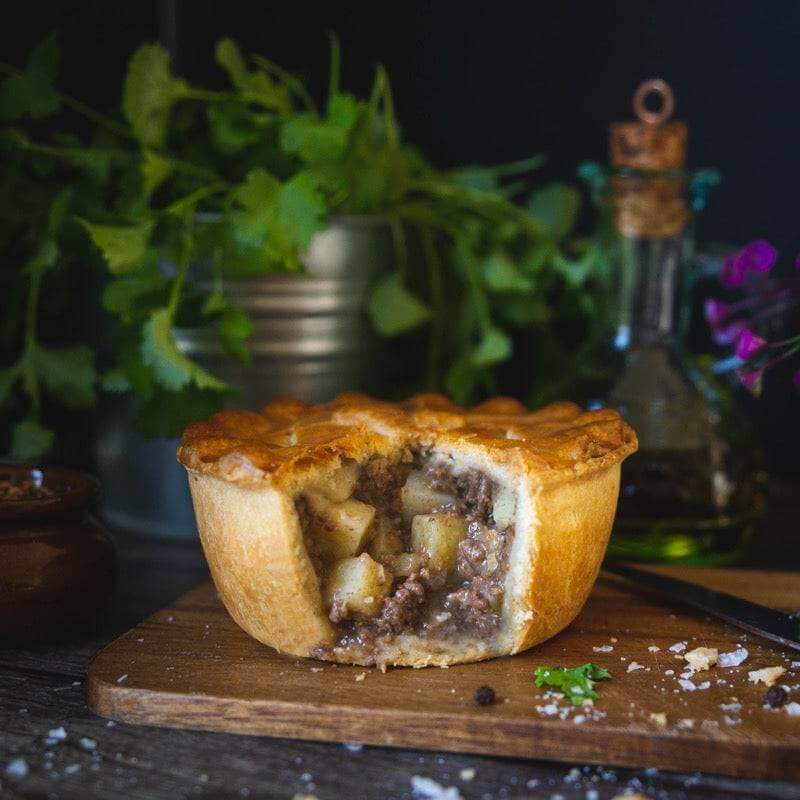 Mud Pies' Hearty Beef and Potato Pie (Beef and Potato Pie), packed with grass-fed steak and rich gravy in golden pastry, sits on a board with herbs nearby.