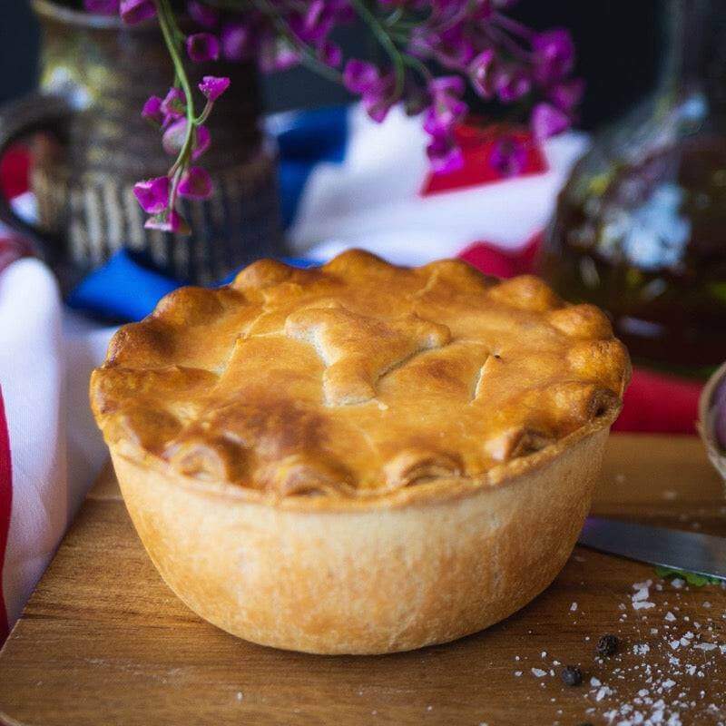 Mud Pies Hearty Beef and Potato Pie (Meat and Potato Pie), golden-brown with crimped edges, rests on wood; purple blooms and tricolour cloth behind.