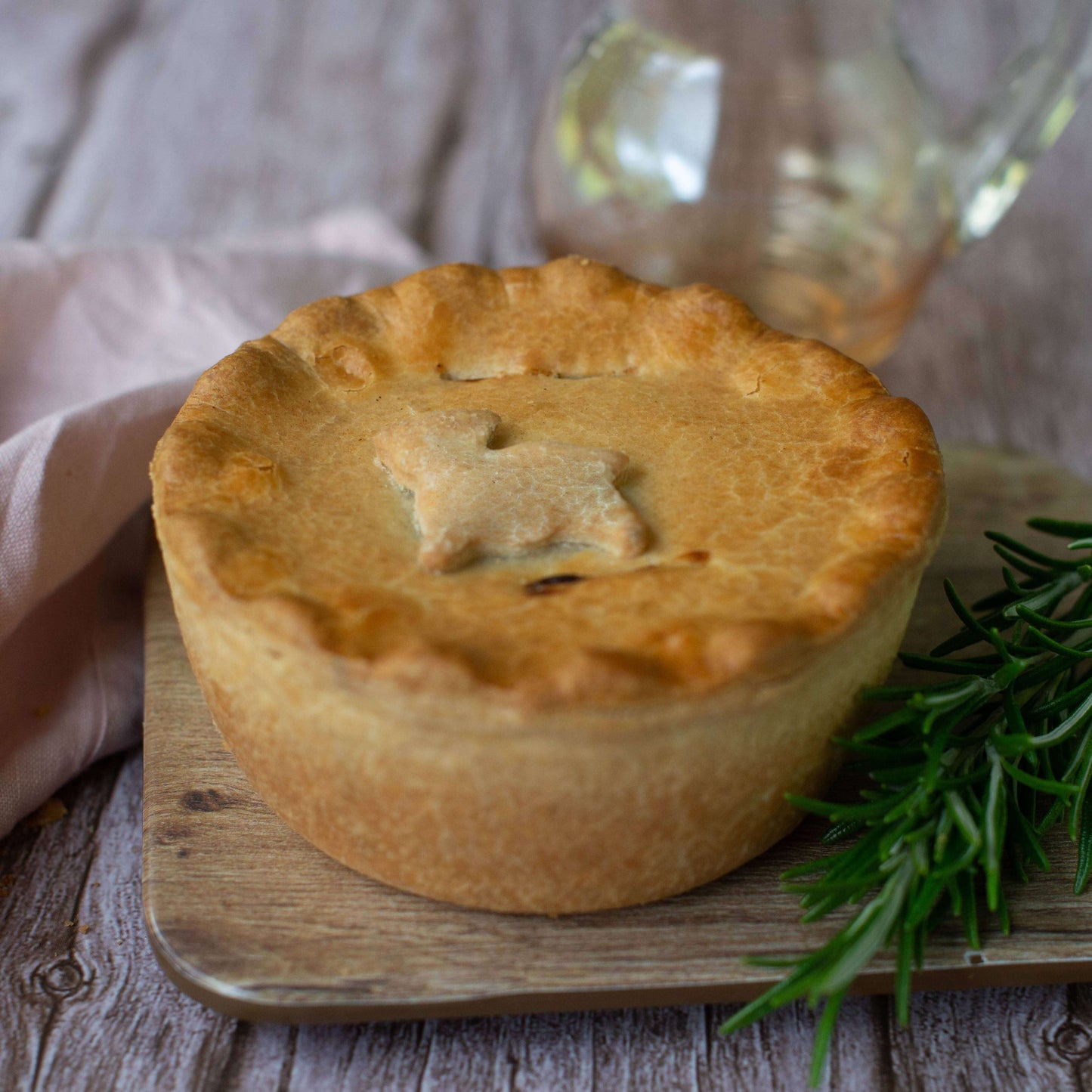 Mud Pies Lamb, Redcurrant and Rosemary Pie, golden-brown with a rabbit pastry cutout, rests on wood with rosemary nearby.