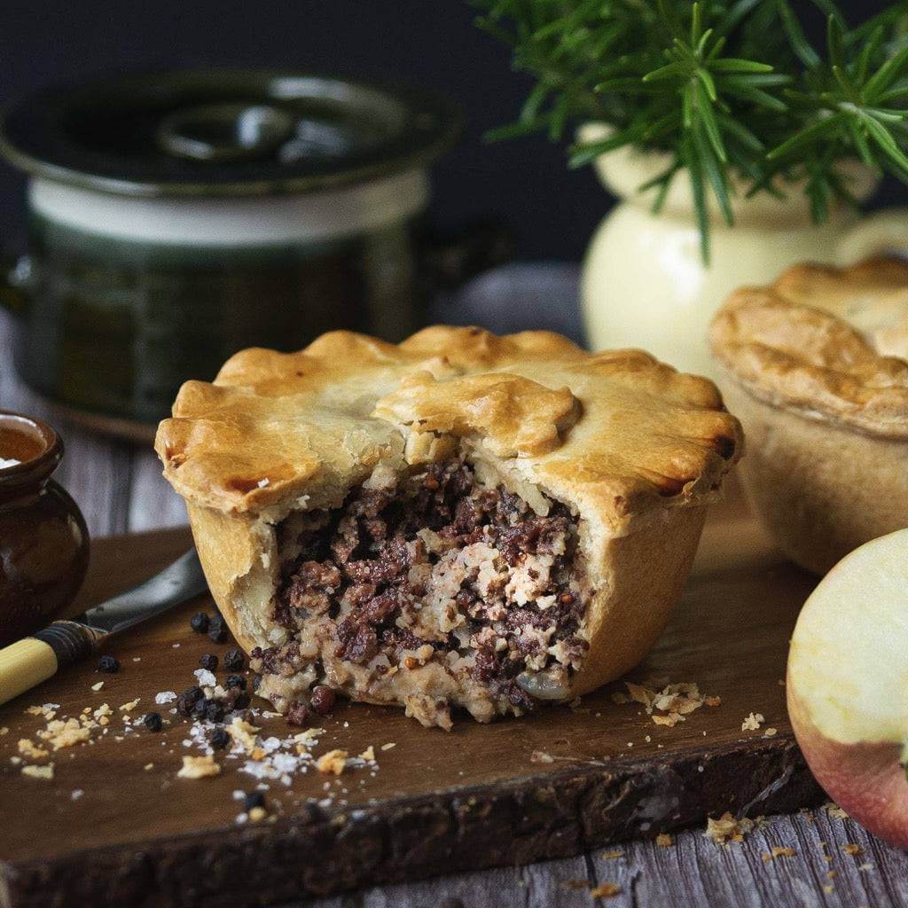 Sausage and Black Pudding Pie by Mud Pies sits on a wooden board, its golden pastry broken to show the rich filling inside.