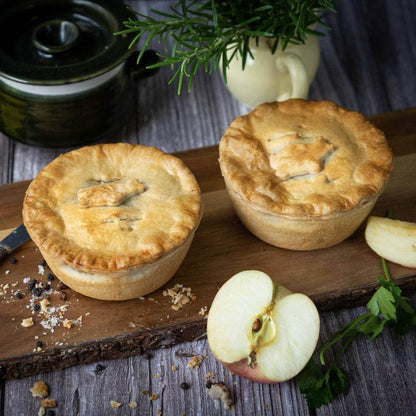 Mud Pies Sausage and Black Pudding Pies, golden-brown, sit on a wooden board with a knife, herbs, peppercorns, and apple nearby.