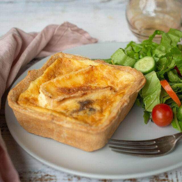 Mud Pies Gluten-Free Red Onion Marmalade and Brie Quiche, with a golden flaky crust, on a white plate next to crisp salad; fork and pink napkin rest on the pale wooden table.