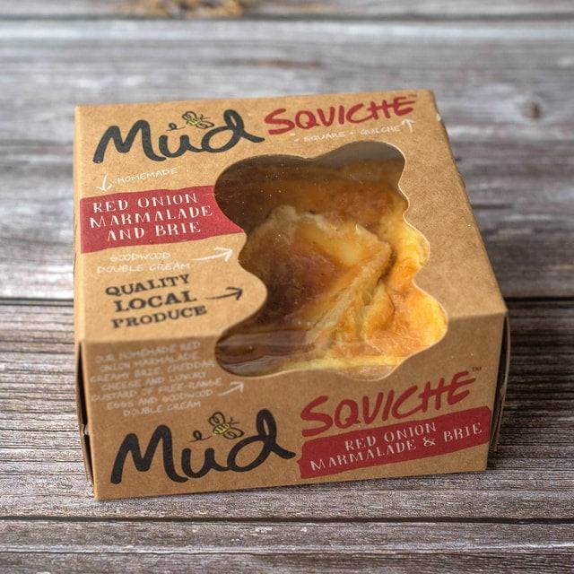 Red Onion Marmalade and Brie Quiche by Mud Pies sits in a square box with a clear window, atop rustic, weathered wooden boards.