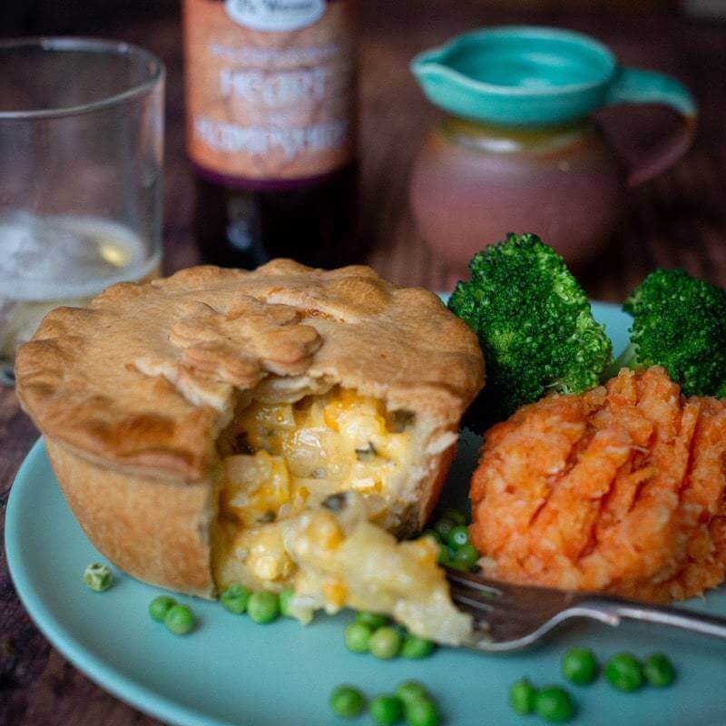 The Mud Pies Gluten-Free Scrumpy Cheese and Onion Pie rests on a plate, its golden crust open to creamy filling, with mash, peas and broccoli.