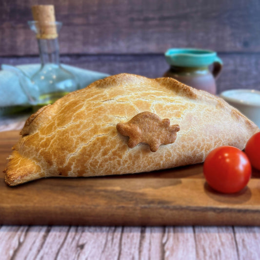 Mud Pies Dinosaur Pasty, a golden-brown Cornish Pasty and crowned with a pastry dinosaur, rests on a wooden board next to two red cherry tomatoes.