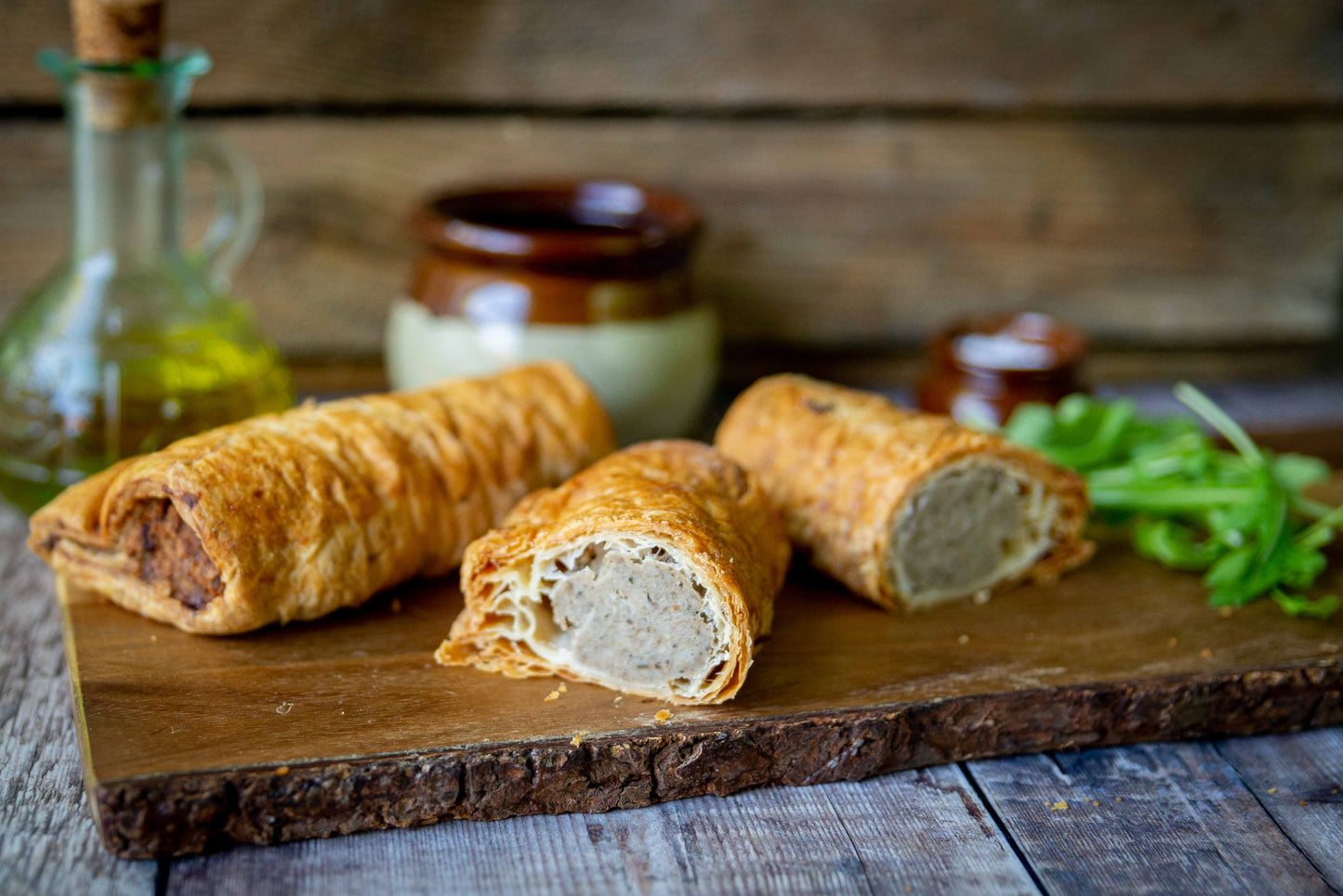 Mud Pies Luxury Sausage Rolls—three golden-brown pastries, one cut open to show the pork filling—on a rustic wooden board, herbs and jars behind.