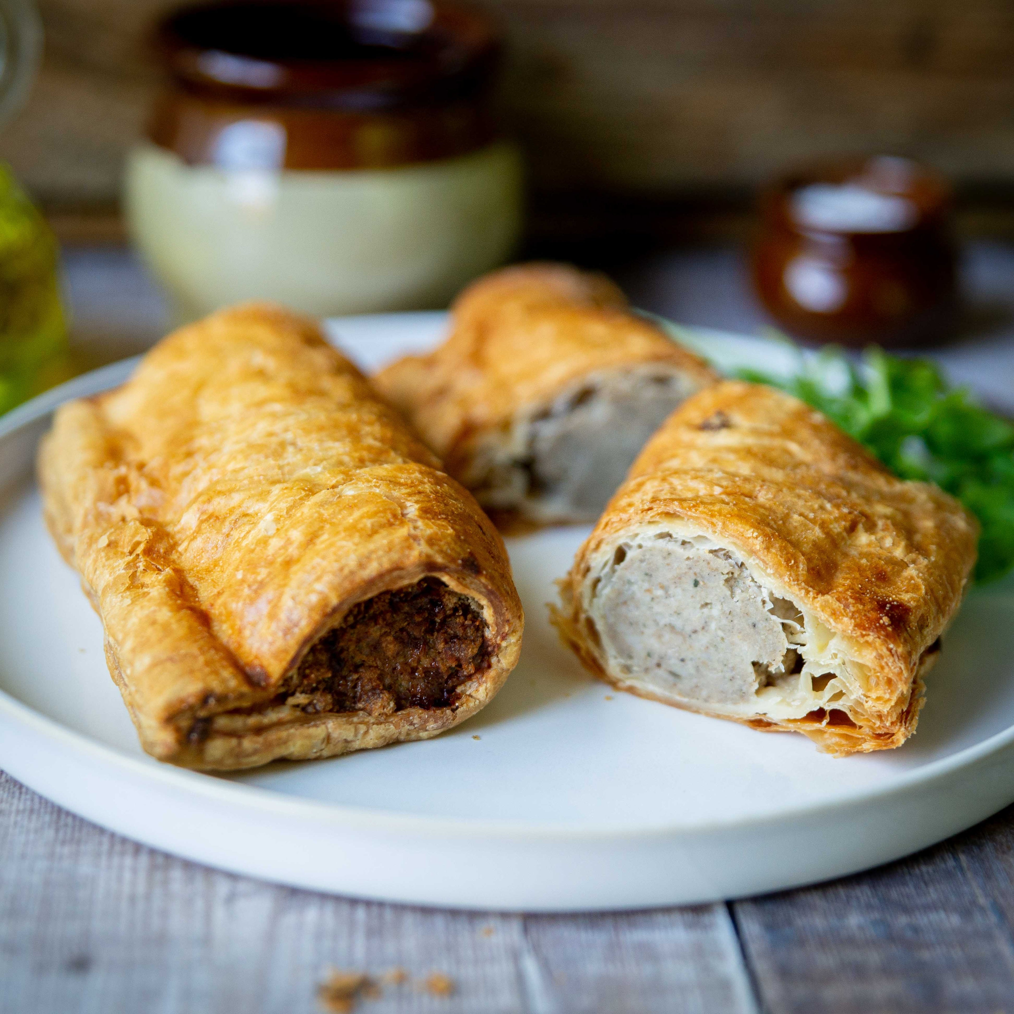Mud Pies Luxury Sausage Rolls, golden and flaky, rest on a white plate—one whole, one sliced to reveal outdoor-reared pork filling.