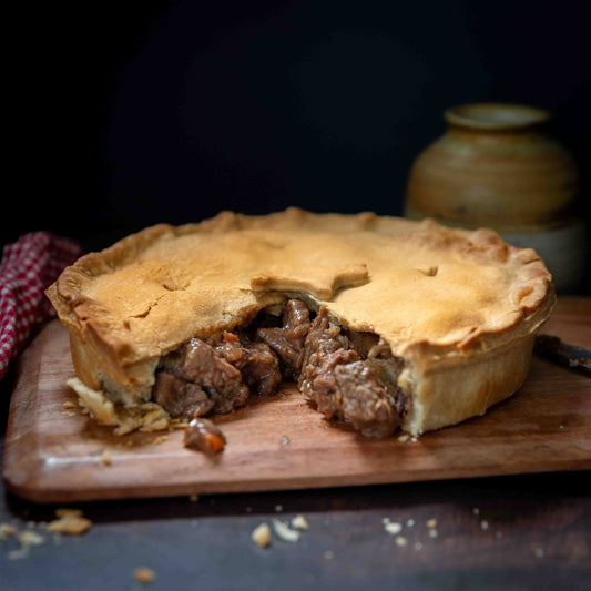The Mud Pies Steak and Ale Family Pie shows golden shortcrust pastry, a cut slice revealing beef in gravy, crumbs on wooden board.