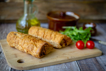 Mud Pies Luxury Sausage Rolls, golden-brown, sit on a wooden board with cherry tomatoes, fresh rocket, oil bottle and rustic pot.