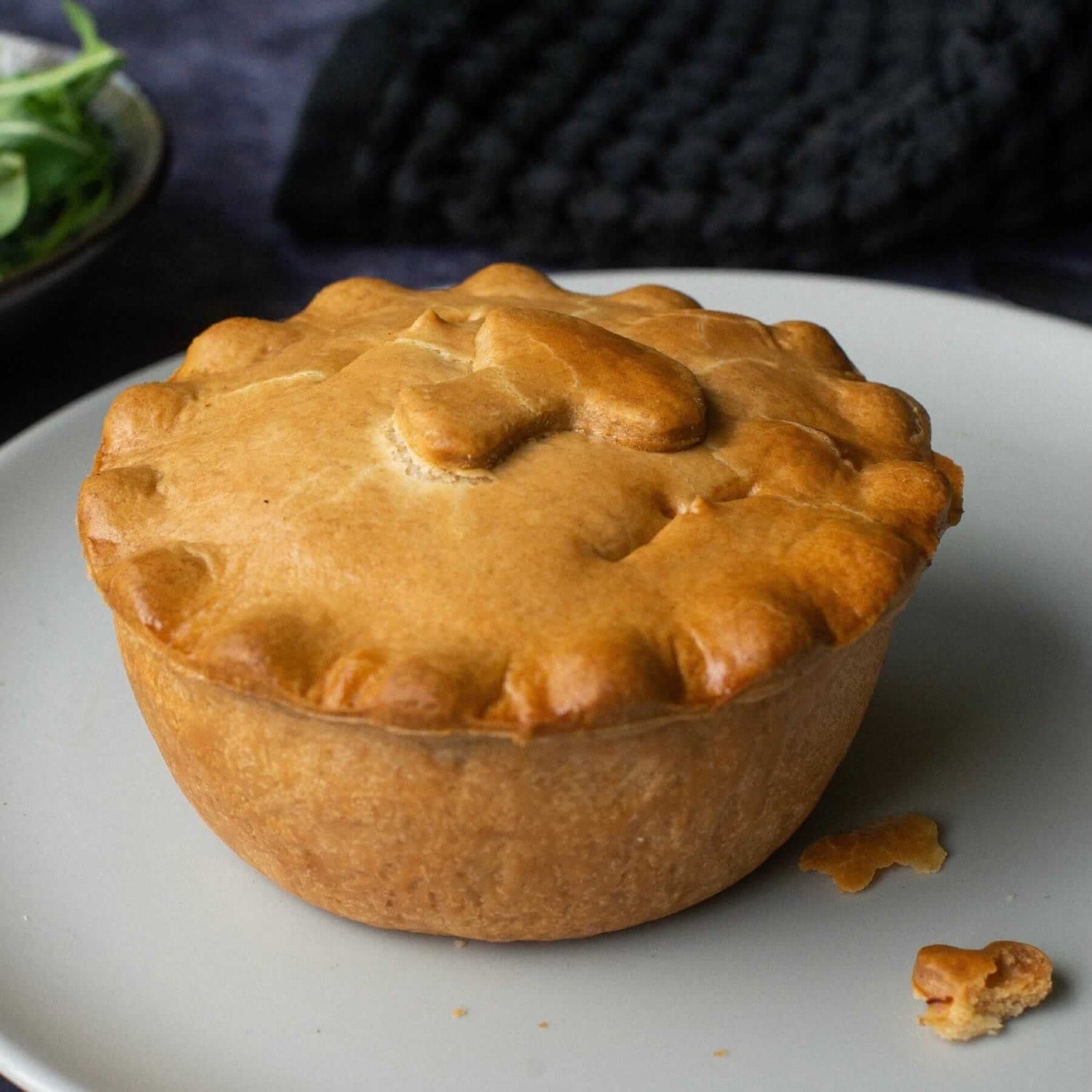 Mud Pies' mushroom and asparagus pie, golden-brown with a fluted crust, sits on a grey plate near green leaves on dark cloth.