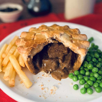 Mud Pies Peppered Steak Pie, golden and half-eaten, reveals rich filling on a white plate with chips and peas over a red tablecloth.
