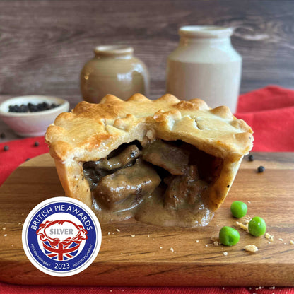 Mud Pies’ Peppered Steak Pie, golden and generously filled, is cut open with green peas, peppercorns, jars and red cloth behind. A 2023 British Pie Awards Silver badge features in the corner.
