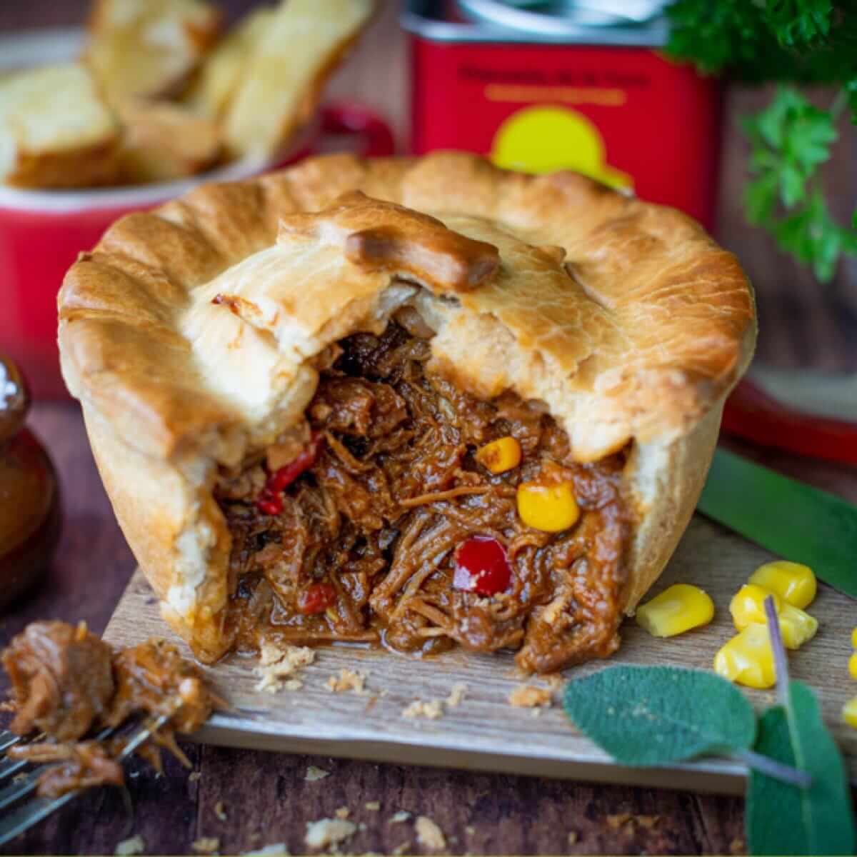 A golden, flaky gourmet pie with shredded beef, corn, and red peppers sits on a board; fork and blurred herbs and red tin behind.