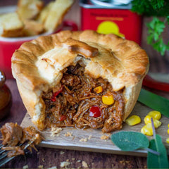 A golden, flaky gourmet pie with shredded beef, corn, and red peppers sits on a board; fork and blurred herbs and red tin behind.