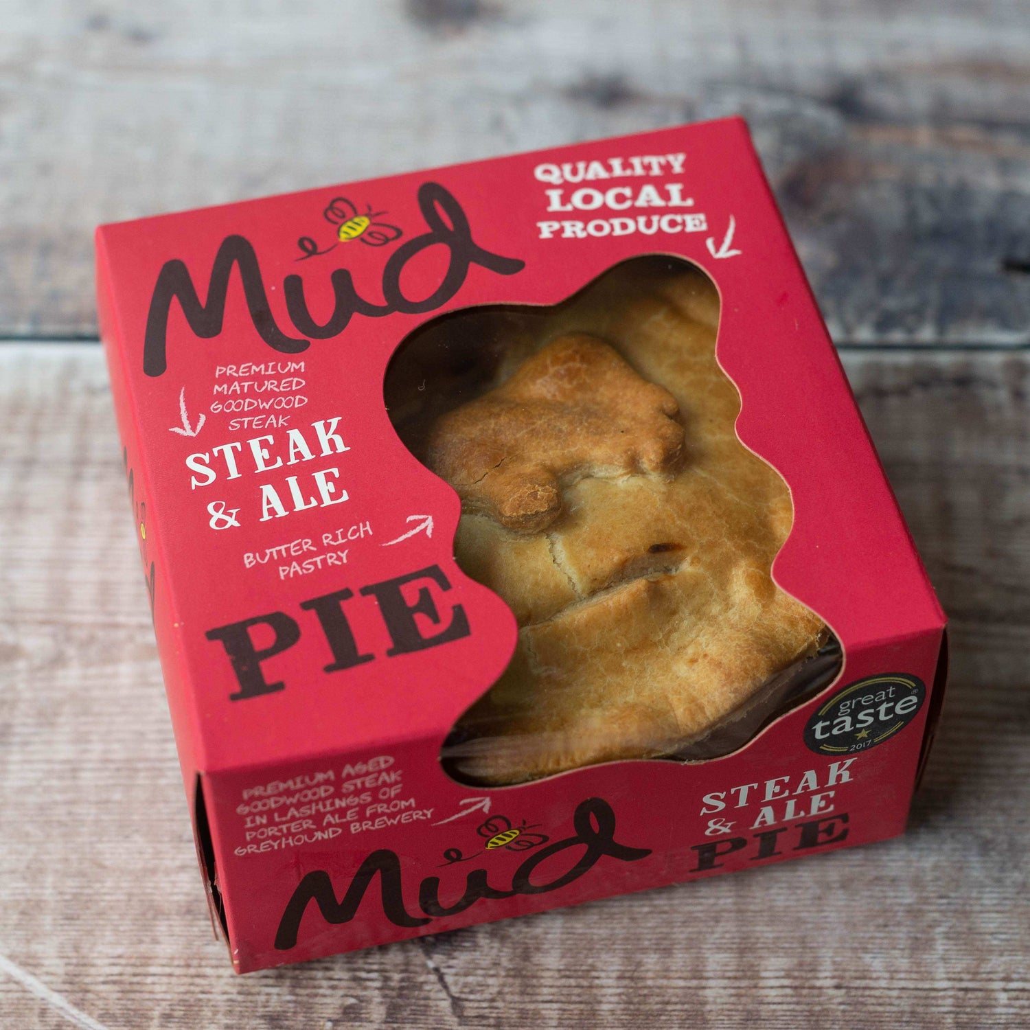 Mud Pies Steak and Ale Pie sits boxed on wood; red packaging shows its name, with a cow-shaped golden pie seen through the window.