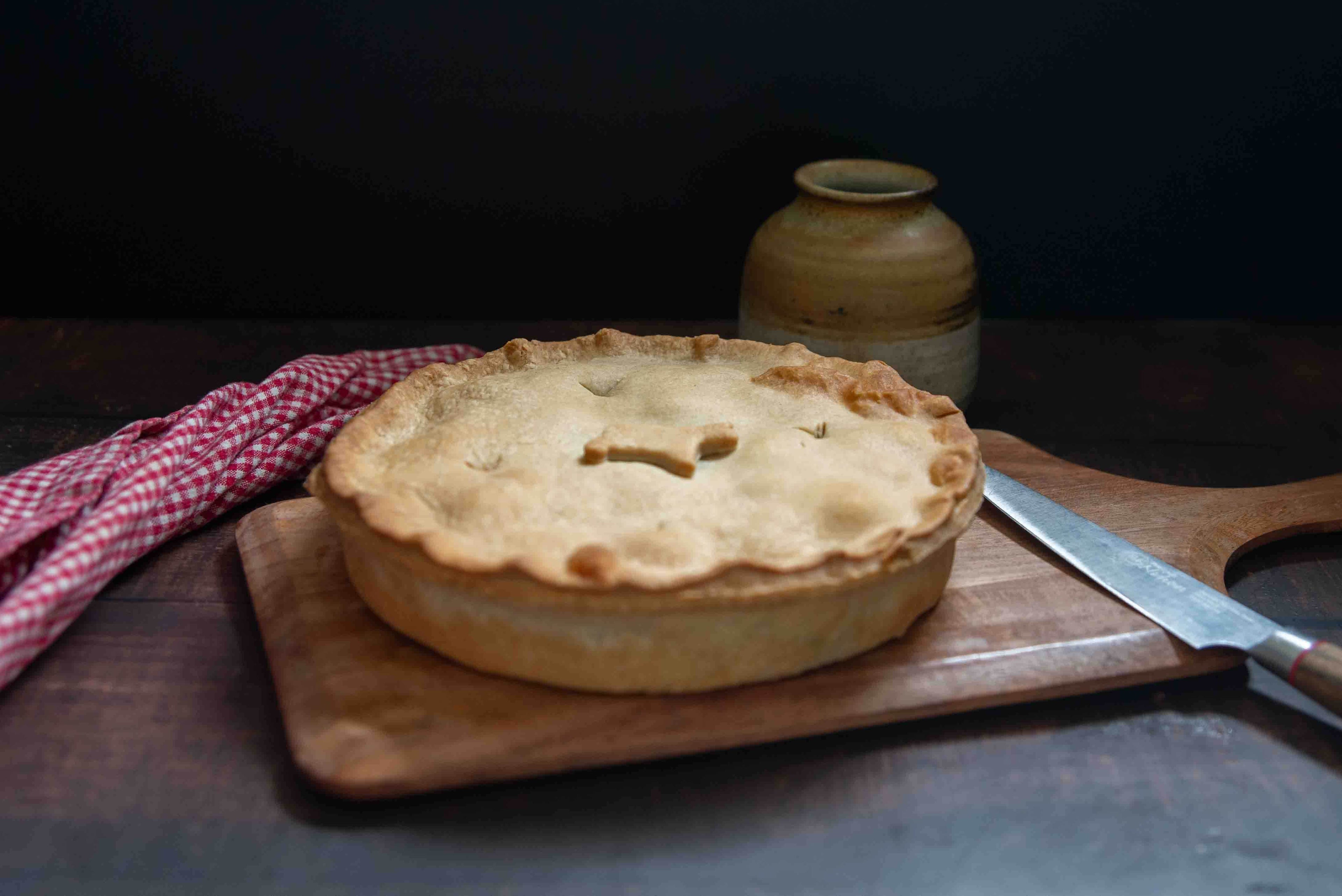 Mud Pies' golden steak and ale family pie sits on a wooden board with knife, checkered cloth, and rustic jug in the dim background.