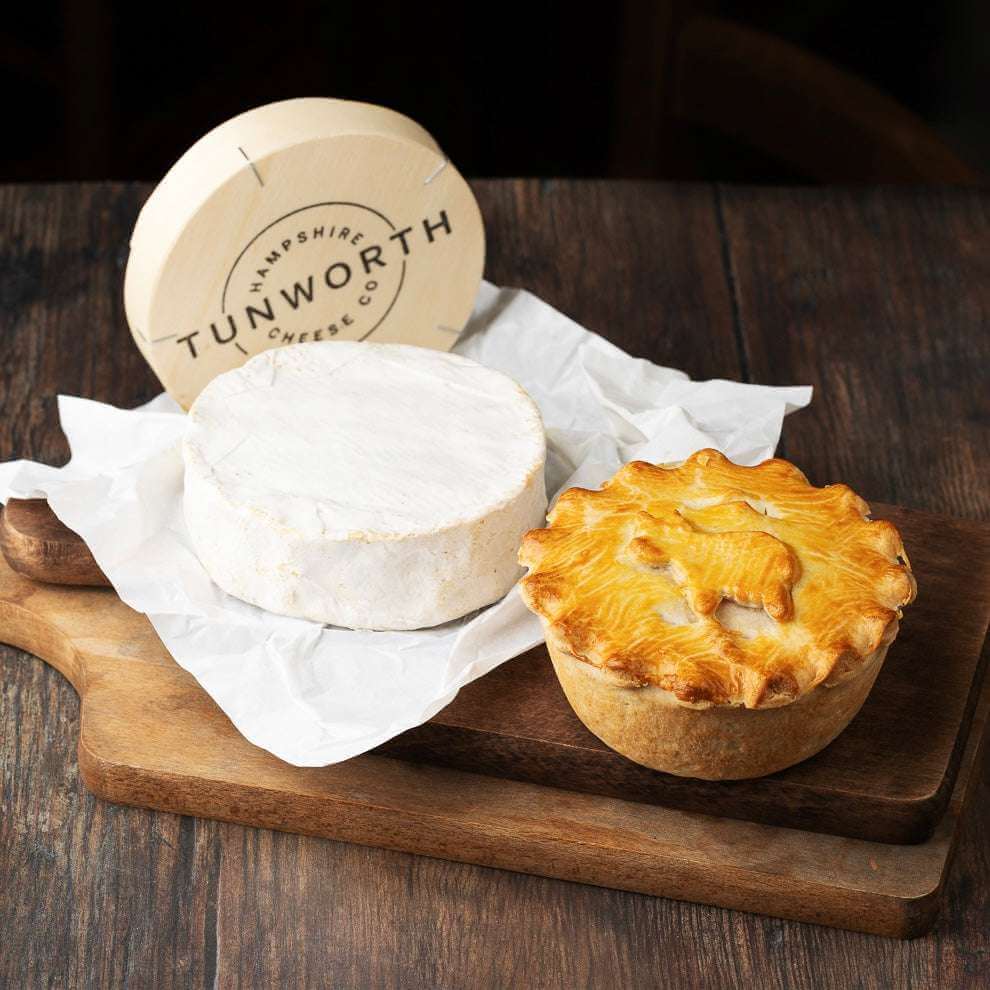 Tunworth cheese sits on parchment beside a golden Mud Pies Steak and Tunworth Cheese Pie, both on a wooden board; the cheese box reads “Tunworth Hampshire Cheese Co.”.