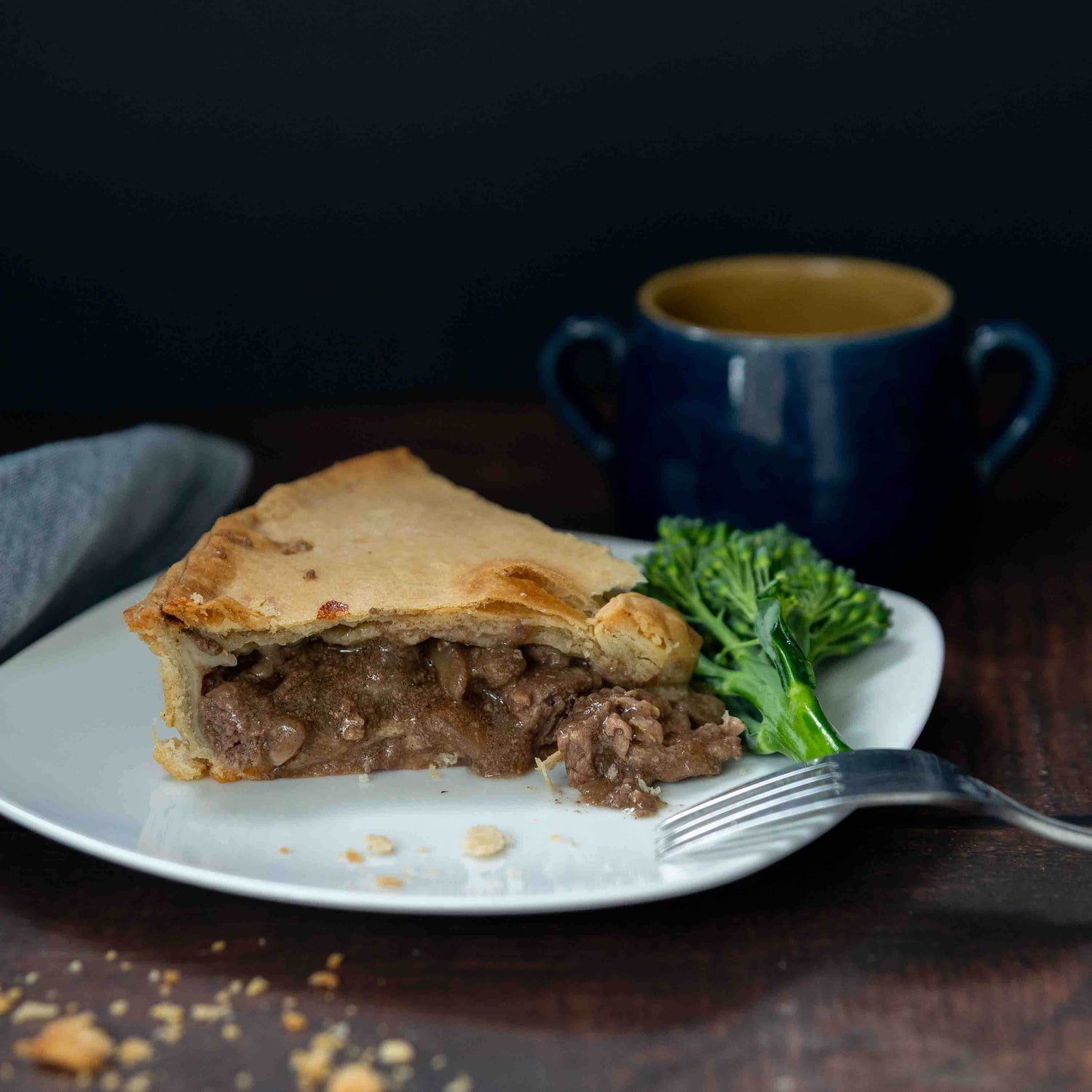 Mud Pies Steak and Kidney Family Pie with golden shortcrust pastry, broccolini, a fork, and blue mug on a dark wooden table.