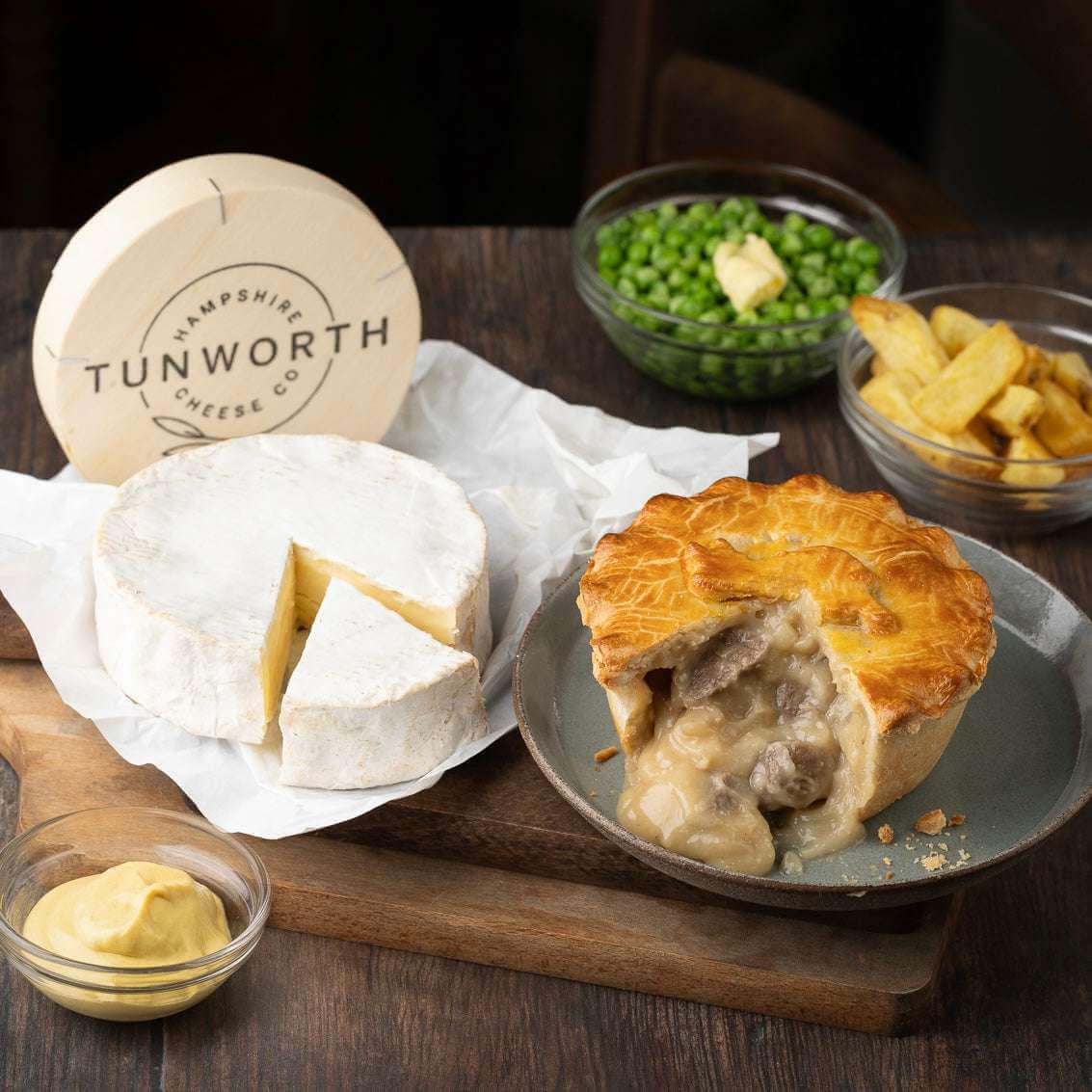 Mud Pies Steak and Tunworth Cheese Pie with a creamy centre on rustic wood, beside sliced cheese, buttered peas, chunky chips, and mustard.