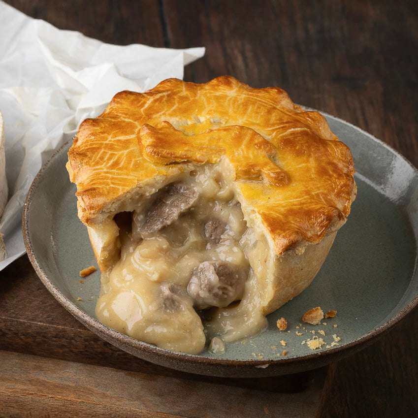 Mud Pies Steak and Tunworth Cheese Pie, golden-brown, rests on a grey plate; creamy steak filling oozes beside rustic wood and paper.