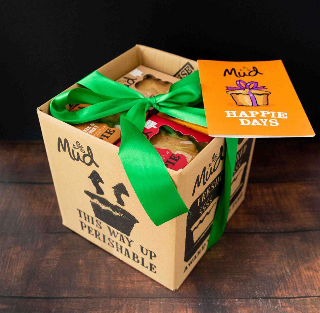 Ultimate Variety Gift Box from Mud Pies, marked "This Way Up Perishable," tied with a green ribbon. An orange card and purple plant nearby.