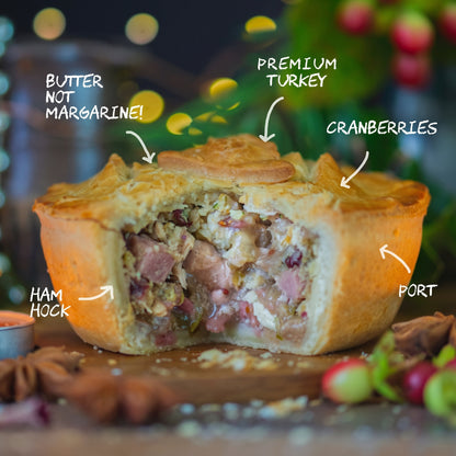 A golden-brown Christmas In A Pie — Turkey and Ham Christmas Pie with a large wedge cut out to reveal a filling of turkey, ham, and cranberries, with text labels pointing to ingredients including butter, premium turkey, cranberries, ham hock, and port.