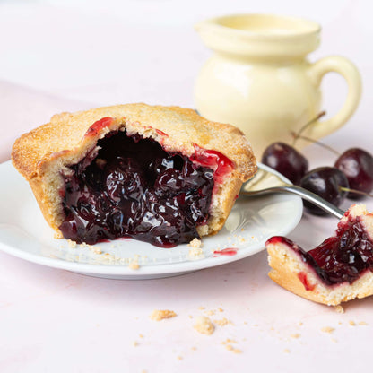 Mud Pies Black Cherry Pie with golden buttery shortcrust pastry on a white plate, cut to show glossy cherries; crumbs nearby, yellow jug with cream and fresh cherries behind on pale pink.