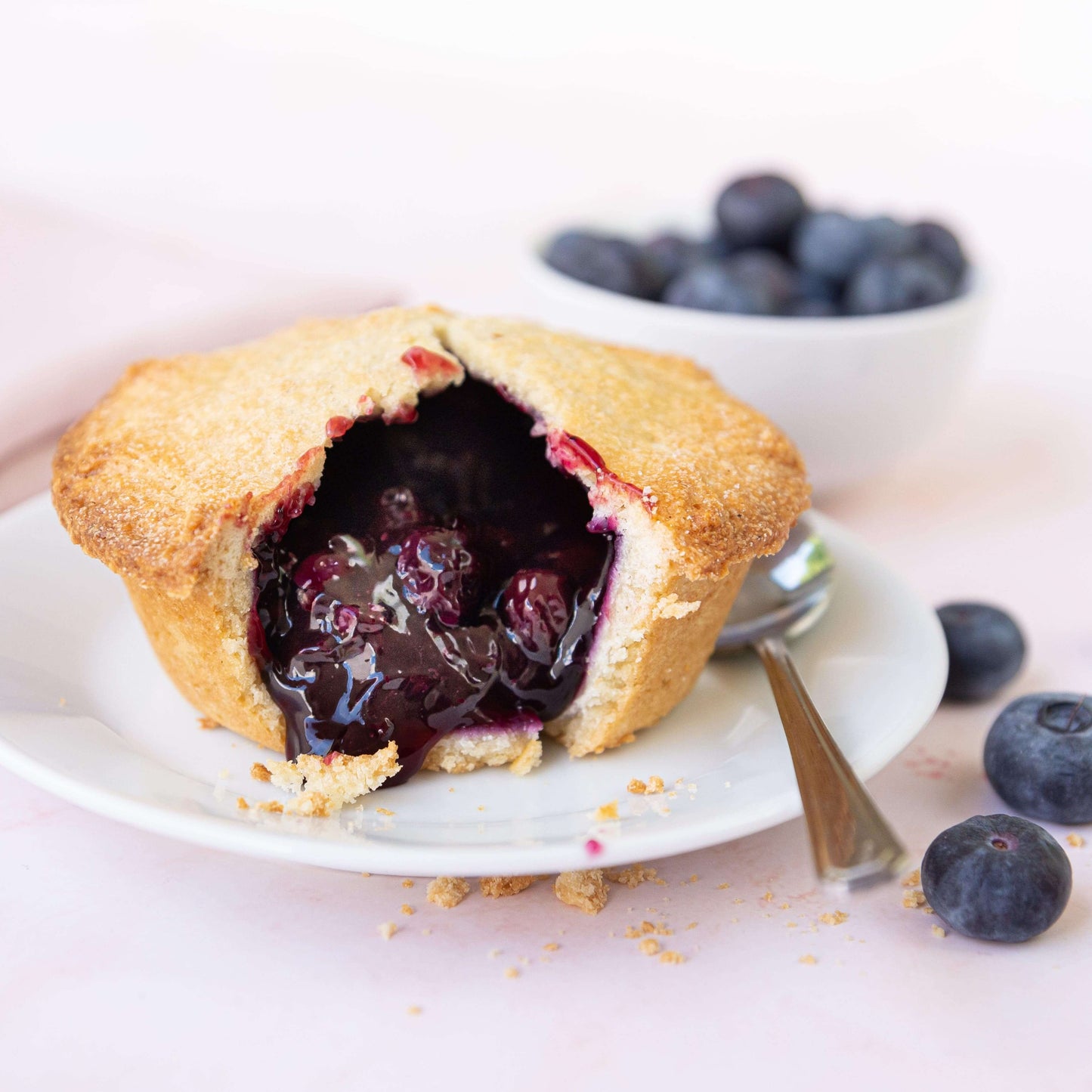 Mud Pies’ Blueberry Pie is a golden mini pie with a cracked top, luscious filling, on a white plate with spoon and fresh berries.