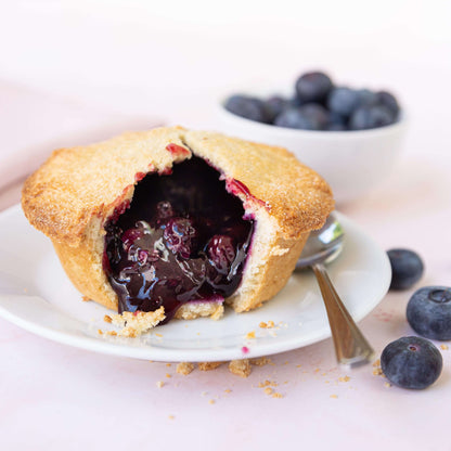 Mud Pies’ Blueberry Pie is a golden mini pie with a cracked top, luscious filling, on a white plate with spoon and fresh berries.