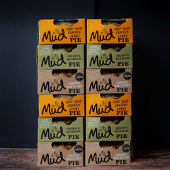Twelve Mud Pies Chicken Pie Box 12 Pack stacked in two neat columns on wood, showing bestselling pies in orange, brown, and green packs.
