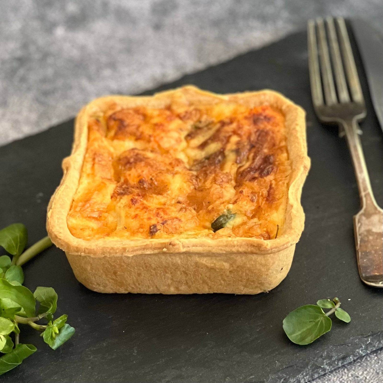 The Mud Pies Hot Smoked Trout Watercress Squiche—a golden-brown square quiche—on slate, herbs, knife and fork nearby.