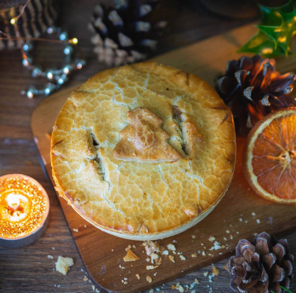 The "Christmas In A Pie" by Mud Pies shows golden shortcrust pastry topped with a tree, set on wood among pinecones and holly.
