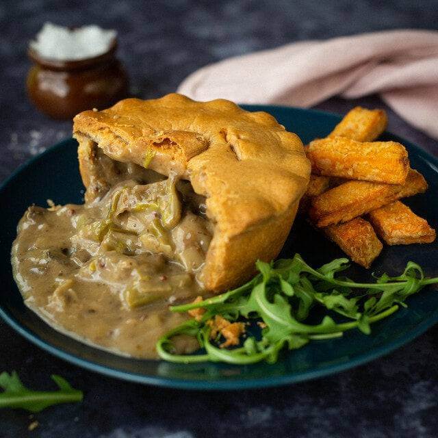 Mud Pies’ Mushroom and Asparagus Pie with golden crust, creamy filling, crispy chips, arugula on dark blue; salt jar, pink napkin.
