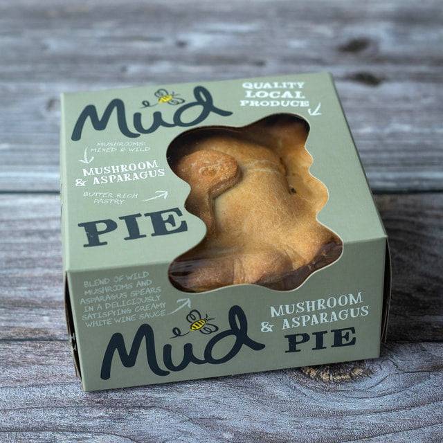 A boxed Mud Pies Mushroom and Asparagus Pie sits on wood. Its pale green box with clear windows reveals the golden, creamy-filled pie.