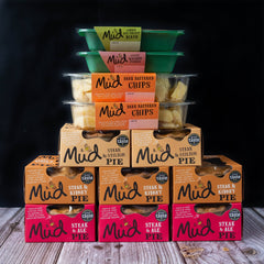 Mud Pies Steak Dinner stacked in a pyramid atop wood, with green trays of carrot-parsnip mash, orange trays of chips, black backdrop.