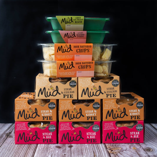 Mud Pies Steak Dinner stacked in a pyramid atop wood, with green trays of carrot-parsnip mash, orange trays of chips, black backdrop.