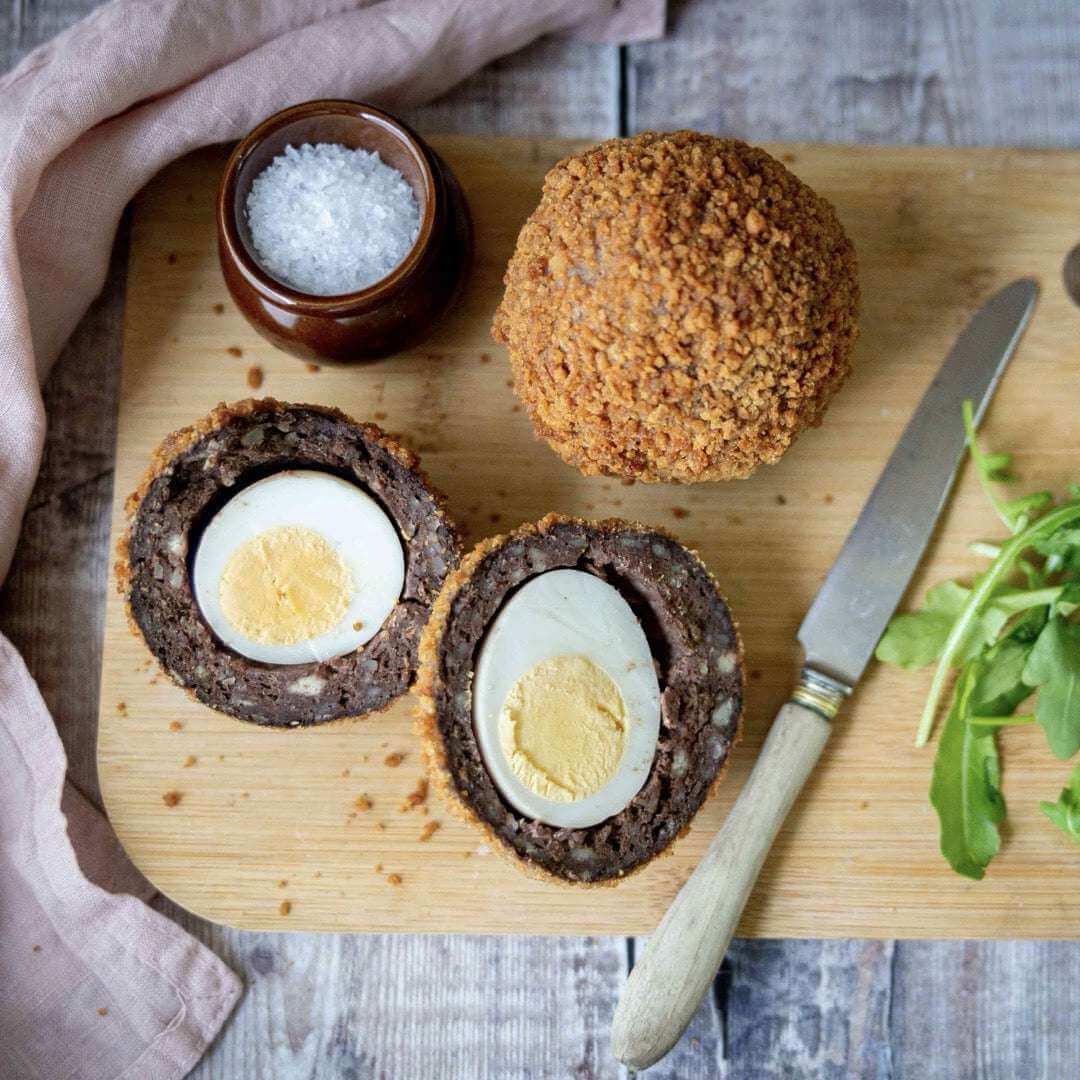Mud Pies Scotch Egg on a wooden board—one whole, one sliced to reveal free-range egg in sausage crumb. Arugula, salt, knife, and pink napkin nearby.