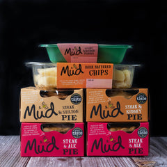 Mud Pies' Steak Dinner meals are stacked neatly on a wooden surface, evoking cosy British comfort against a dark backdrop.