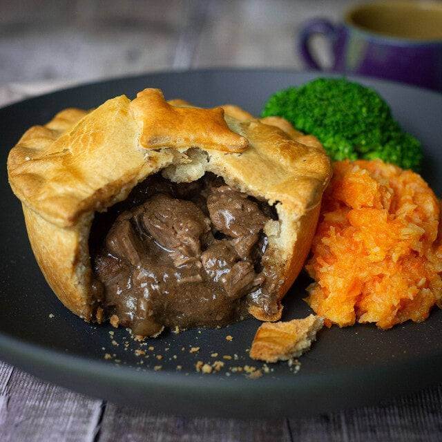 Mud Pies’ Best Pie Box—Dinner for 8 with golden steak and chicken pie, star crust, mashed carrots, broccoli; blurred mug behind.